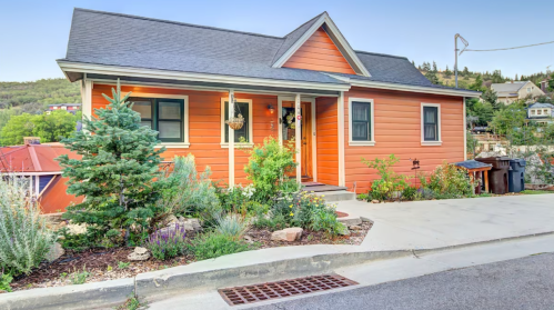 A charming orange house with a sloped driveway, surrounded by greenery and flowers, set against a hillside backdrop.